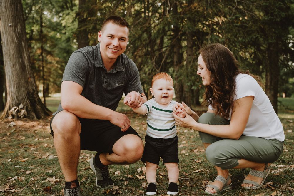 Happy family with parents helping their laughing red-haired baby take first steps in a lush green park.