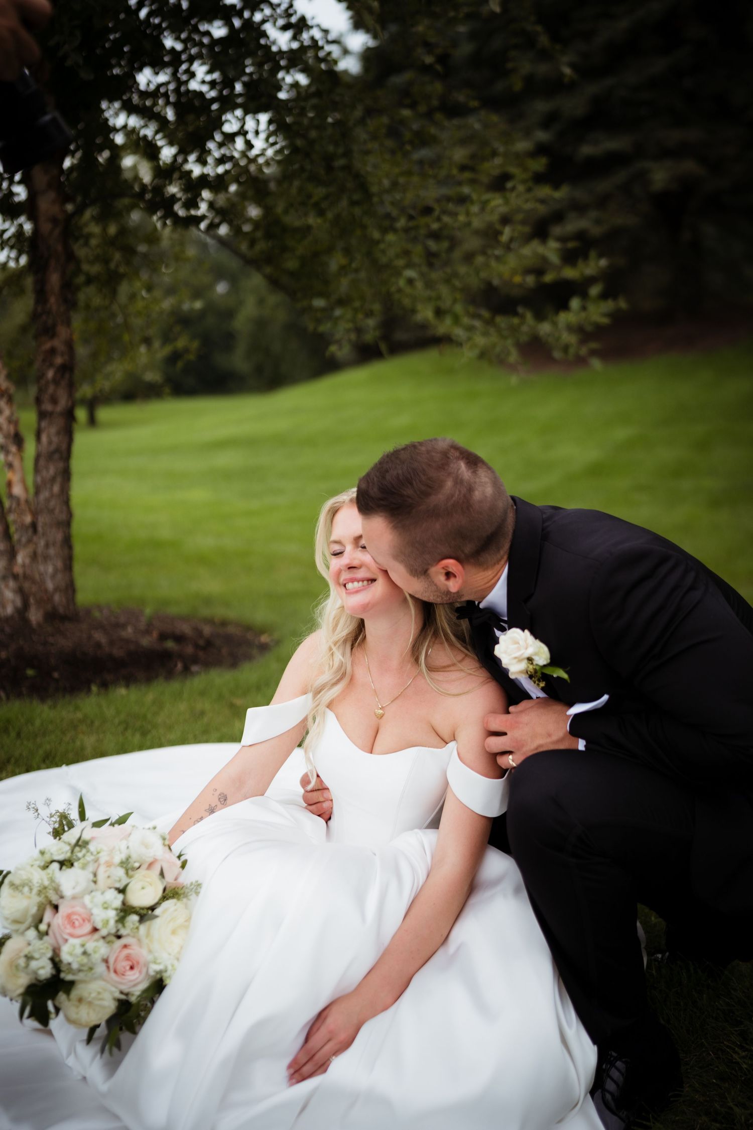 Groom in black tuxedo kisses bride in white off-shoulder gown outdoors, bride holding pink and white bouquet.