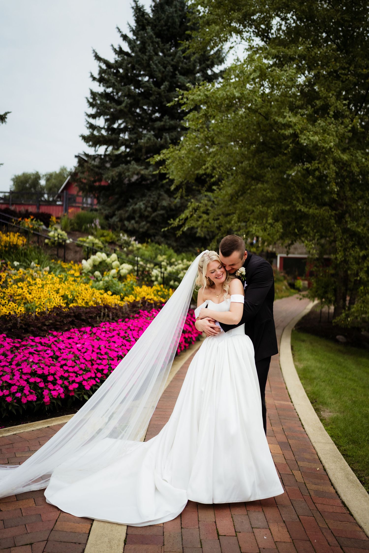 Bride and groom embracing on a brick path surrounded by vibrant flower gardens and lush green trees on their wedding day.