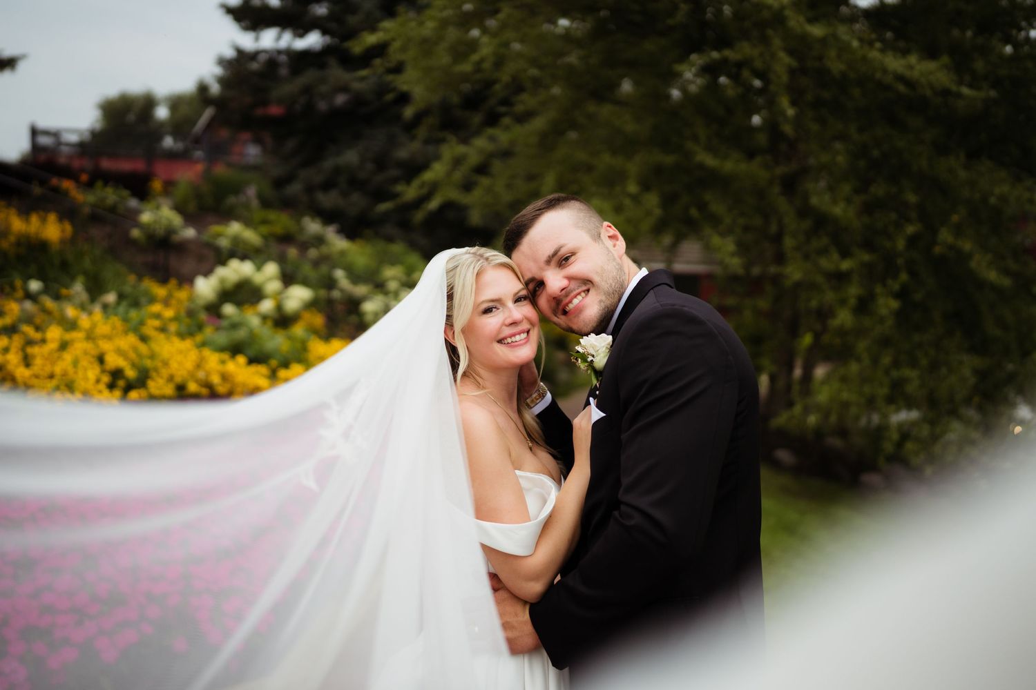 Bride and groom embracing outdoors with flowing veil, colorful garden flowers and greenery in background.