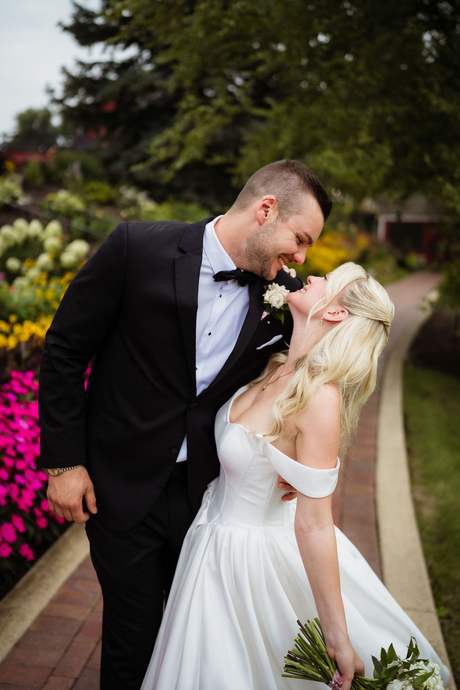 A bride and groom share a romantic moment in a garden, she in a white off-shoulder gown, he in a black tuxedo.