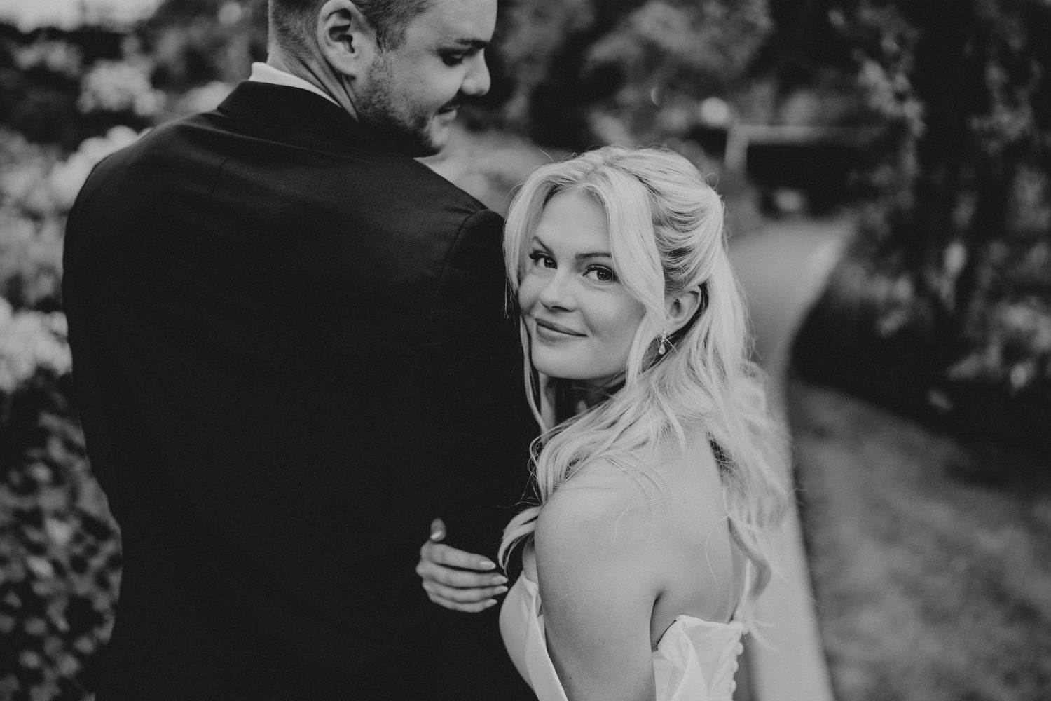 Black and white wedding photo of bride and groom embracing outdoors, bride smiling over her shoulder.