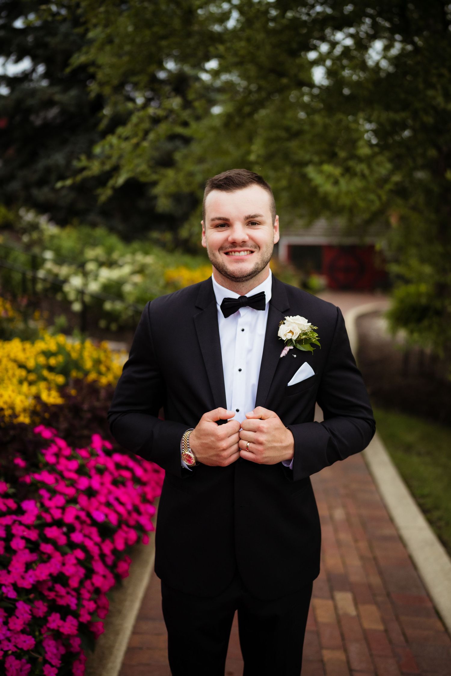 Smiling groom in black tuxedo with boutonniere adjusting jacket in colorful garden setting.
