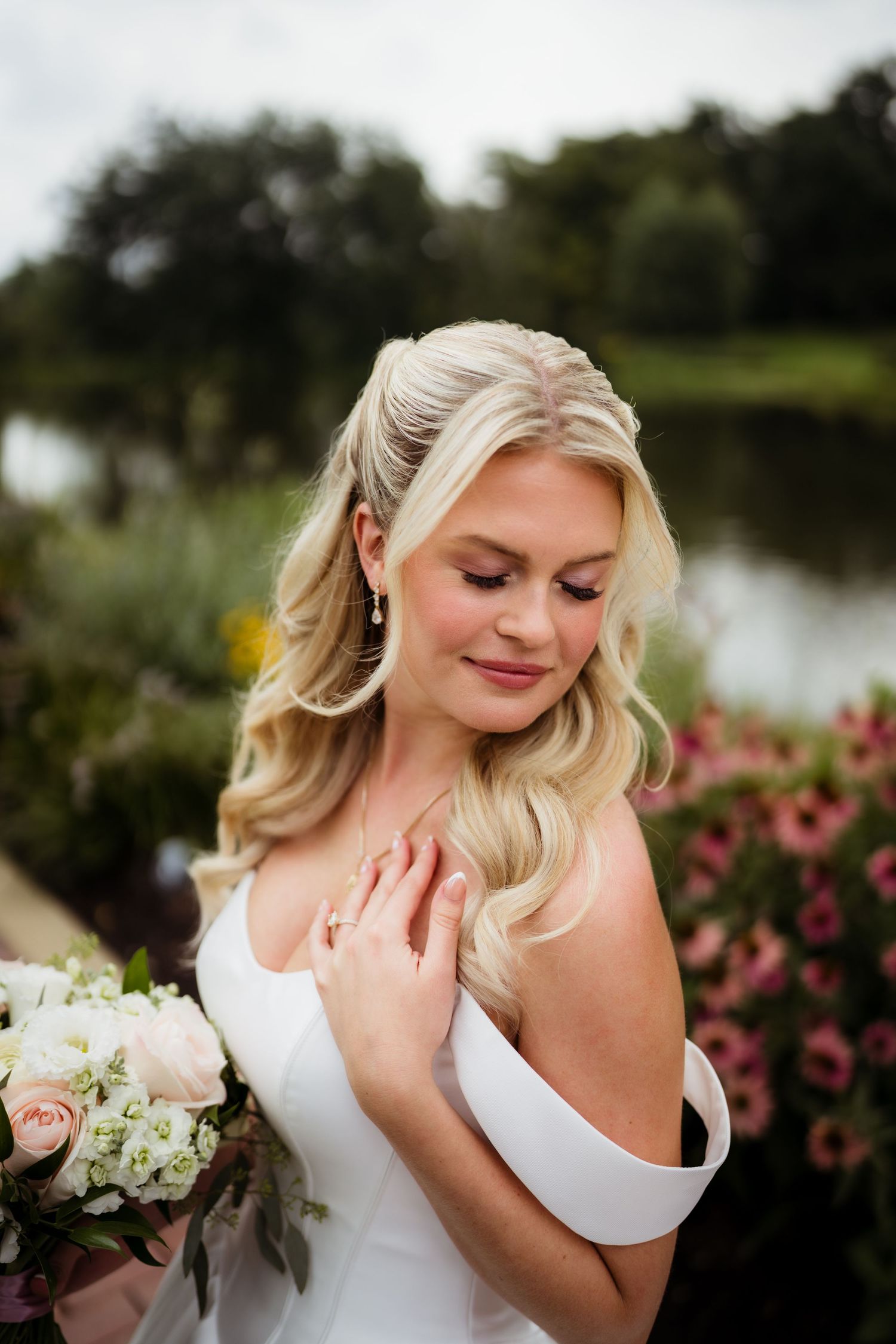 Blonde bride in off-shoulder white gown holds blush bouquet, smiling softly outdoors near a pond with pink flowers.