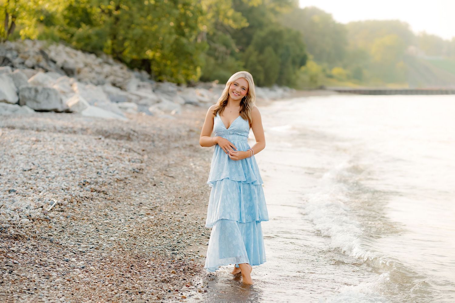 A young blonde woman in a light blue maxi dress stands on a rocky shoreline with gentle waves at her feet.