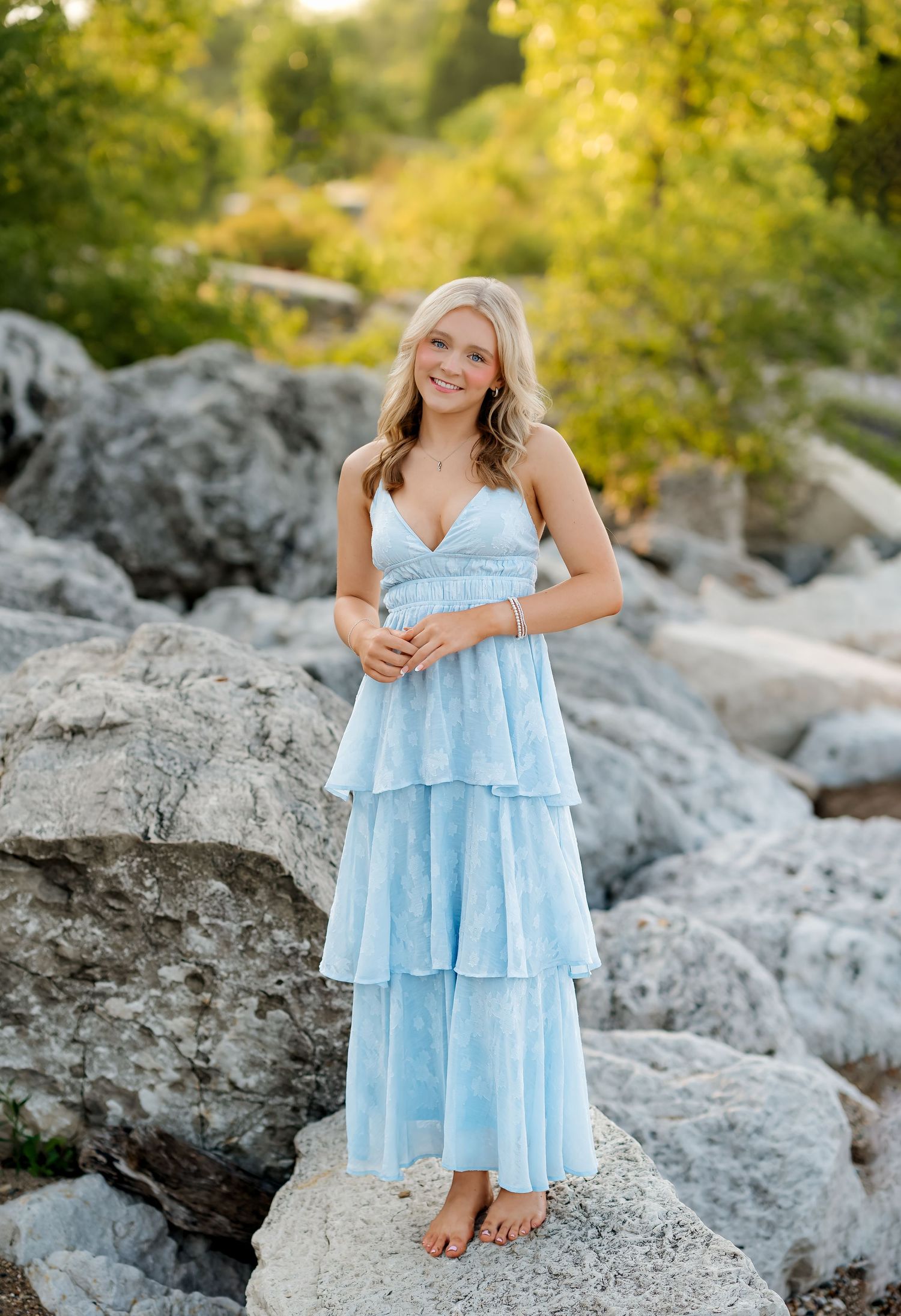 Young blonde woman smiling in a light blue tiered maxi dress, standing barefoot on rocks with greenery background.