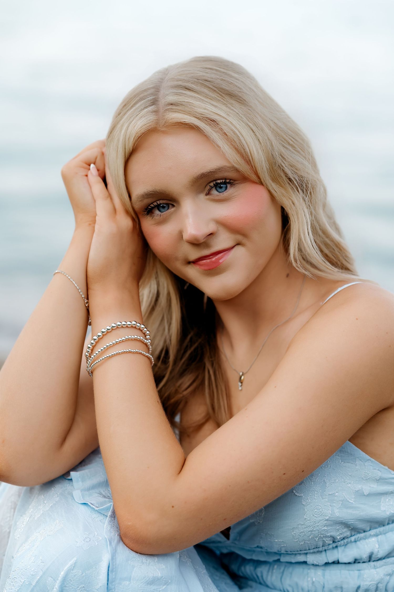 Young blonde woman in blue dress with silver bracelets poses near water for senior portrait photo.