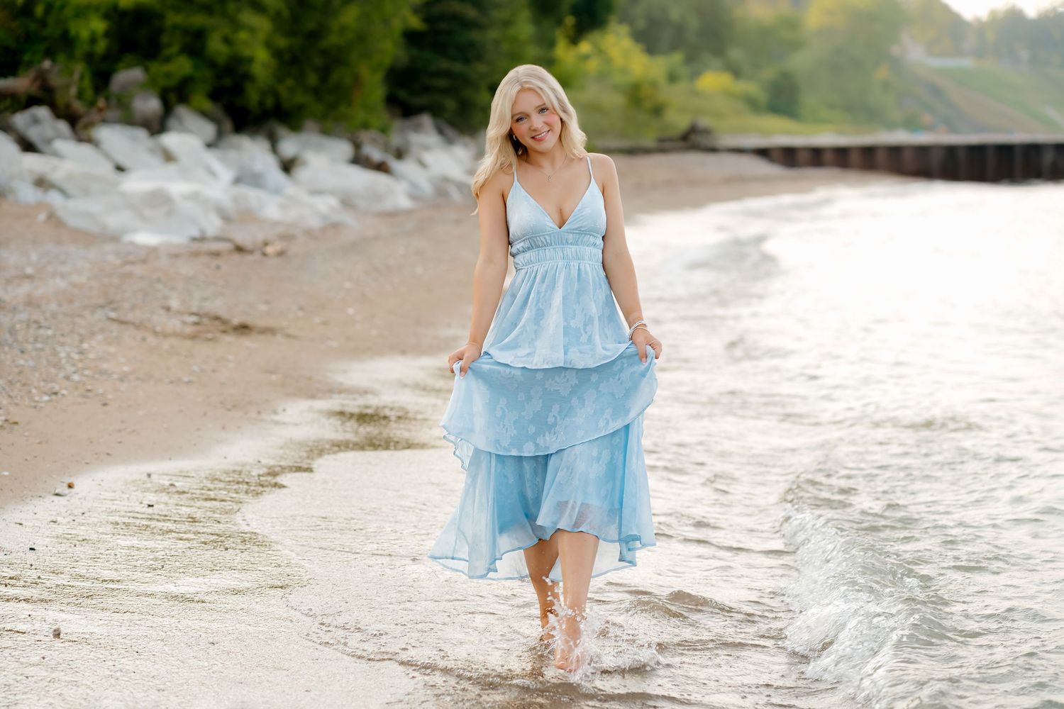 Blonde woman in a light blue sundress walking barefoot along a sandy beach shoreline with gentle waves.