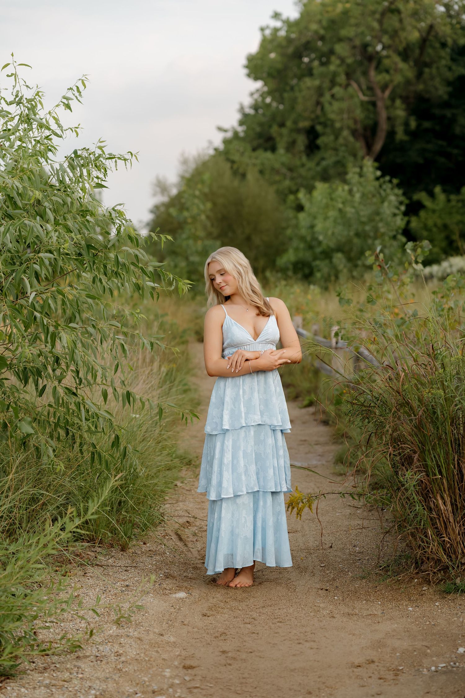 Blonde woman in a light blue tiered maxi dress standing on a dirt path surrounded by lush green foliage.