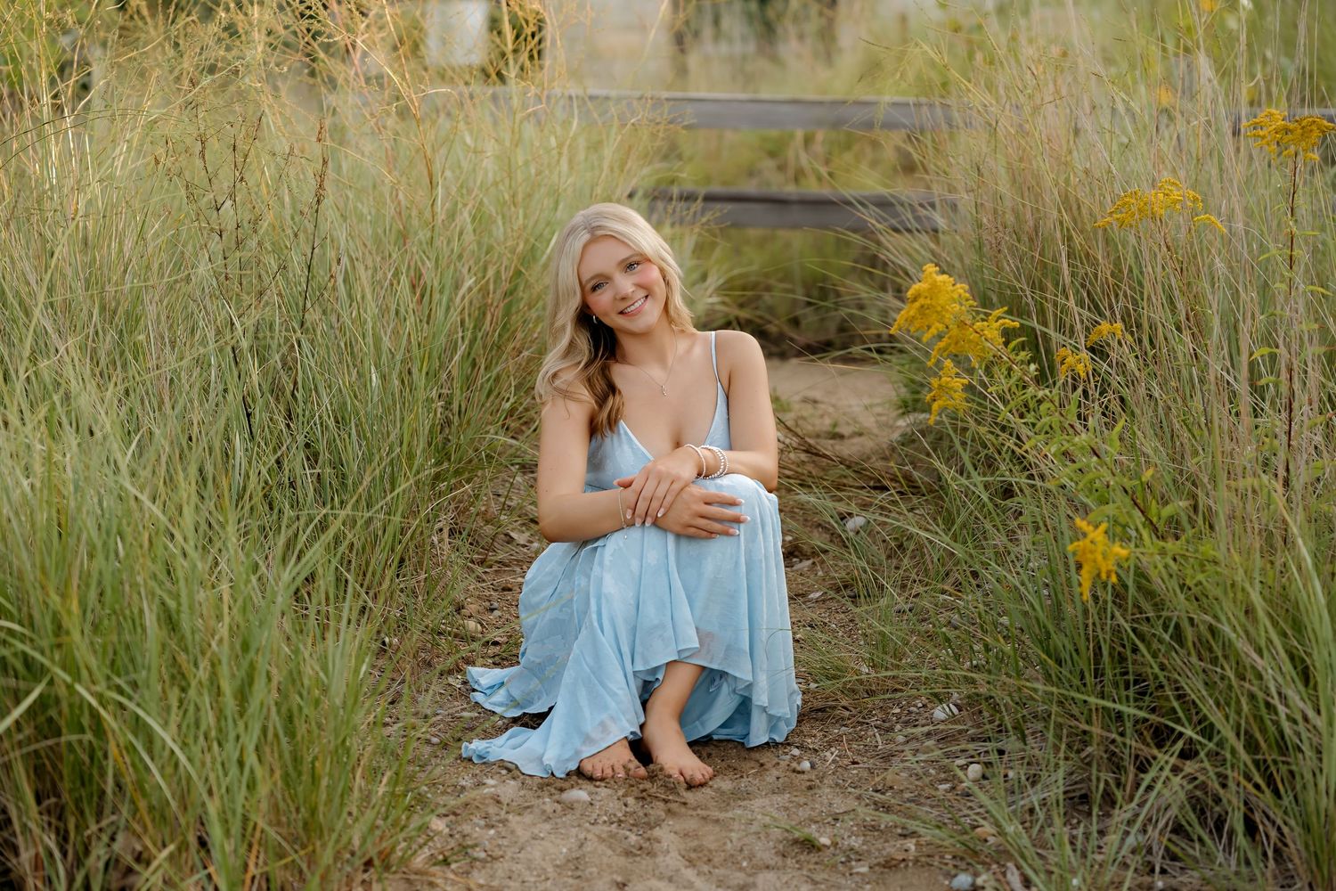 Smiling blonde woman in light blue dress sitting barefoot among tall grass and yellow wildflowers outdoors.