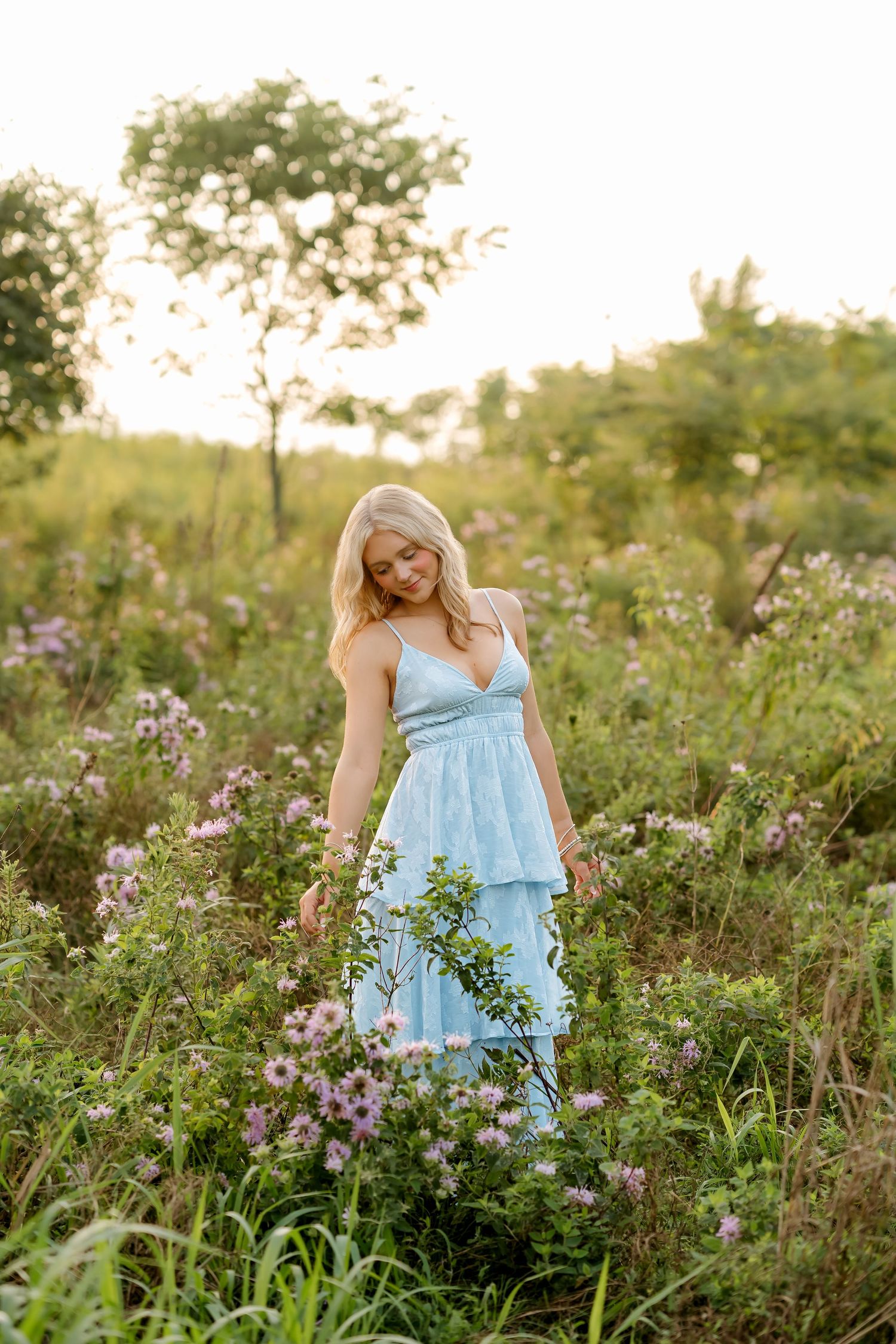 Blonde woman in light blue tiered maxi dress standing among wildflowers in a sunlit meadow at golden hour.