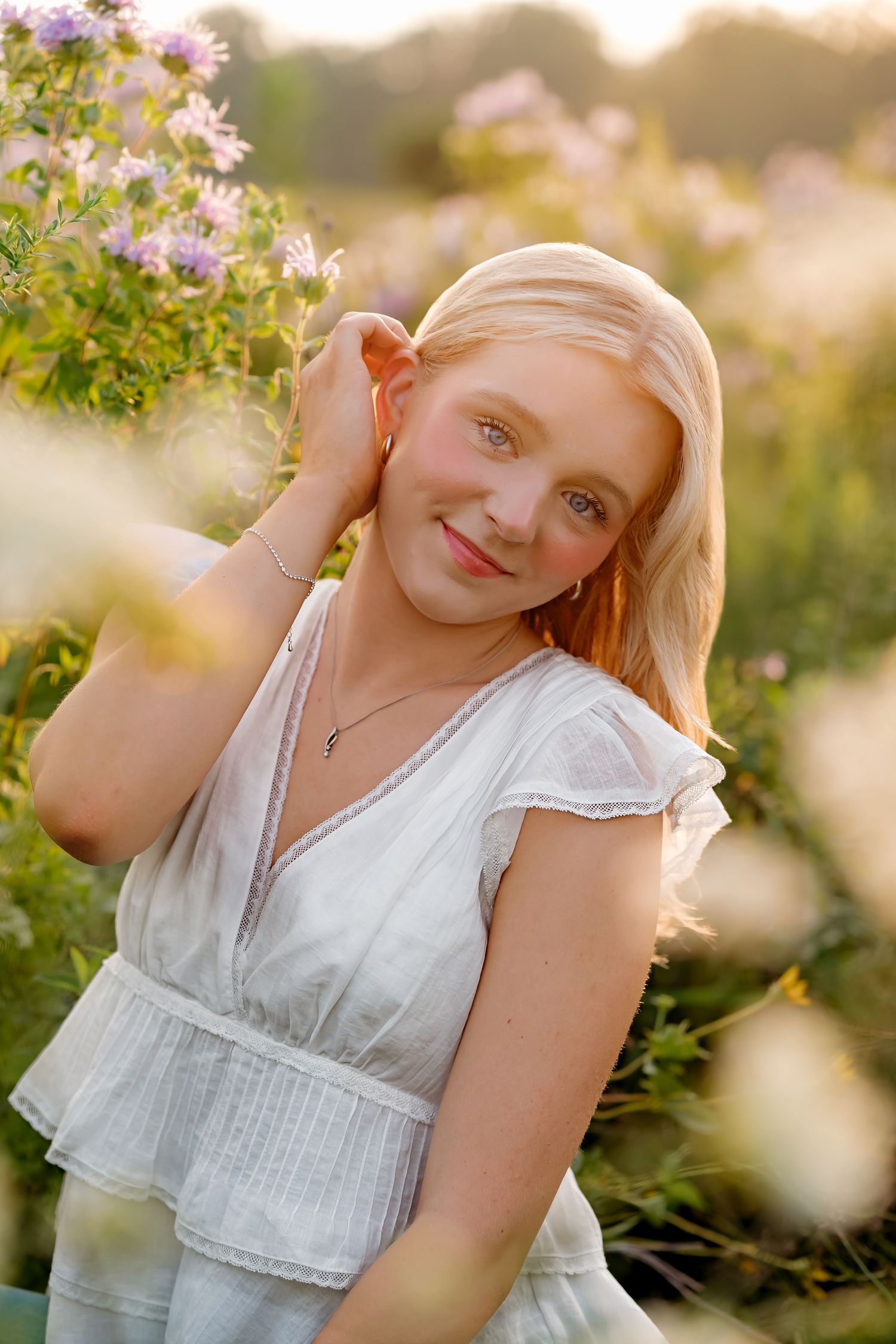 A young blonde girl in a white dress poses among wildflowers in golden hour sunlight, smiling softly at the camera.