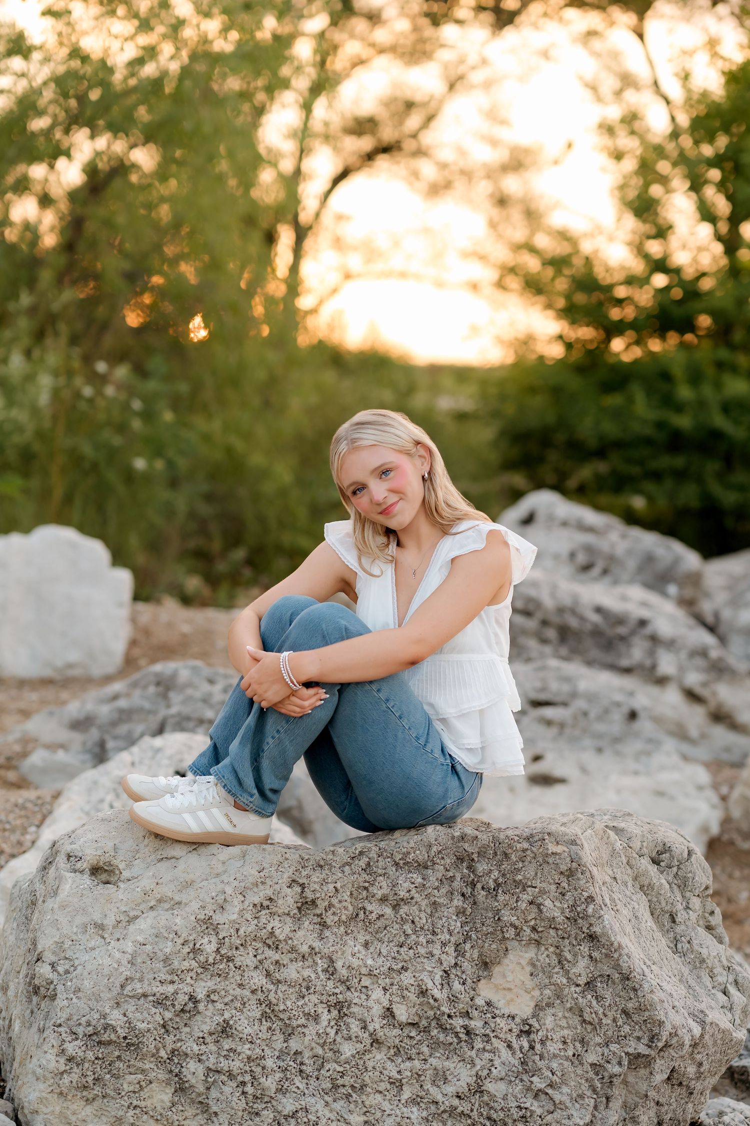 Blonde girl sitting on rocks in white top and jeans, smiling at golden hour with green trees in background.