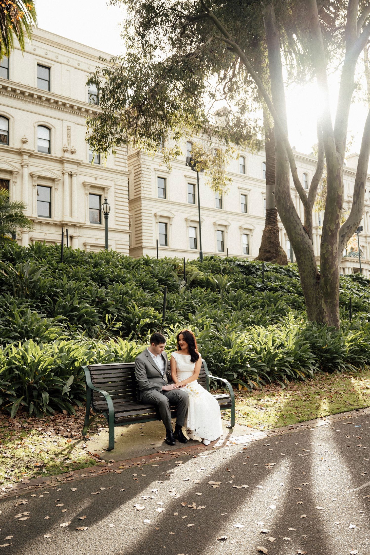 Bride and groom sitting on a park bench surrounded by lush greenery with a grand historic building in the background.