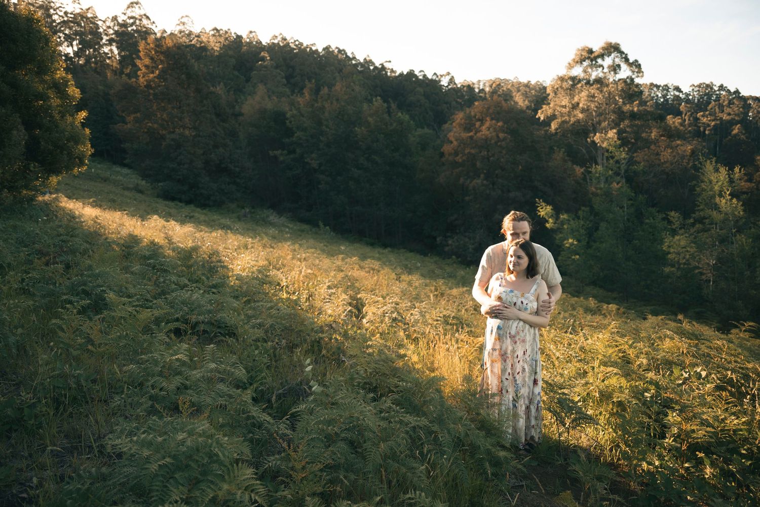 Couple embracing in a sunlit meadow surrounded by lush green forest trees during golden hour outdoors.