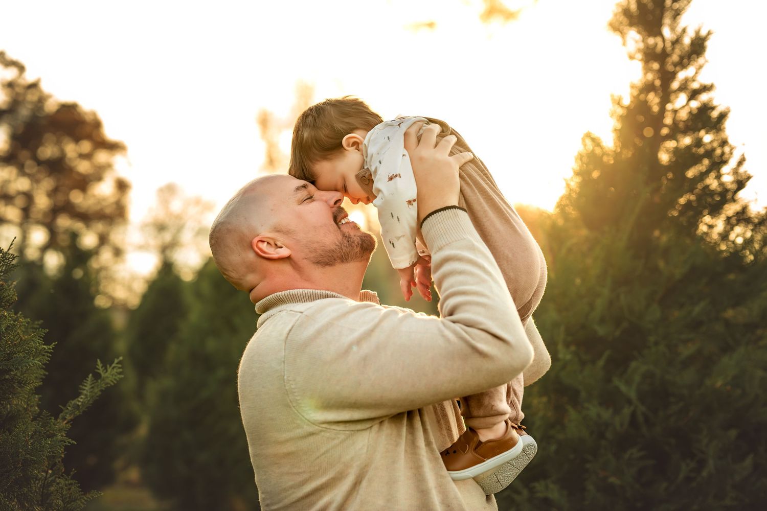 A bald father lifts his baby up to kiss them outdoors among green trees in warm golden sunset light.