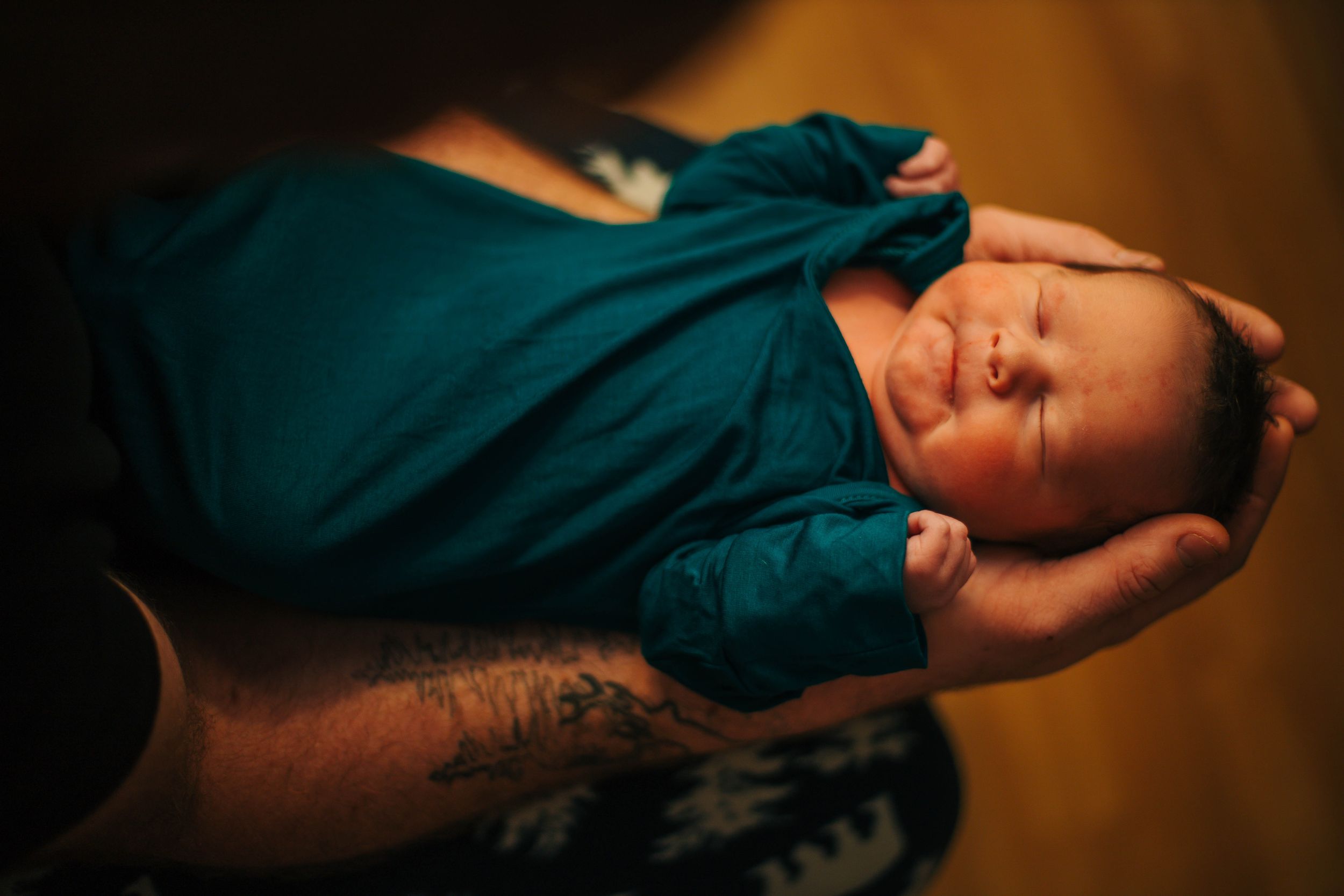 Newborn baby swaddled in teal blanket sleeping peacefully in tattooed parent's hands.