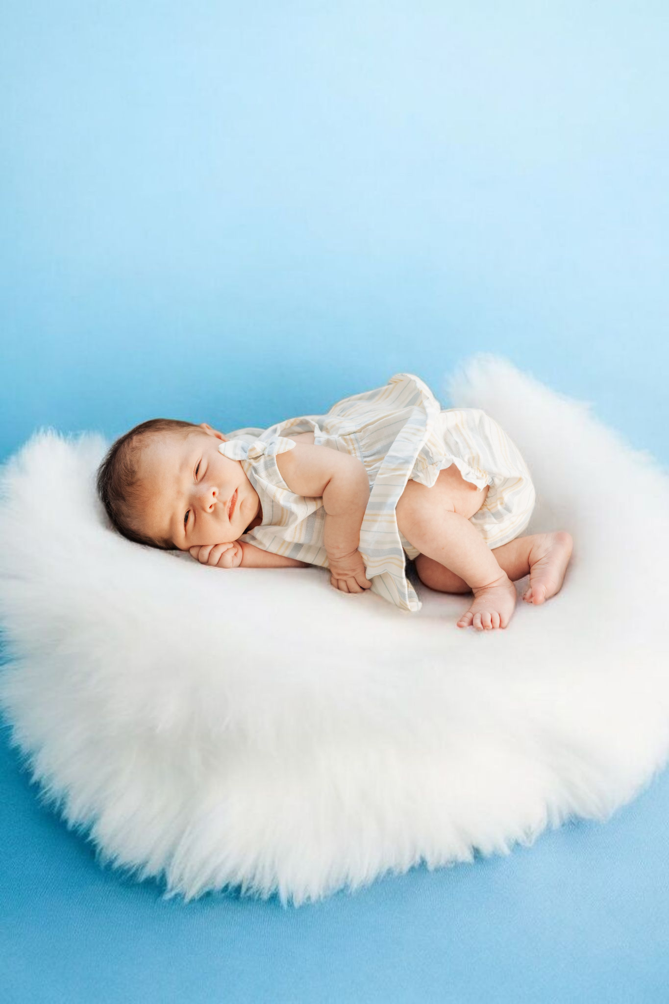Newborn baby sleeping peacefully on white fluffy cushion wearing cream romper against light blue background.