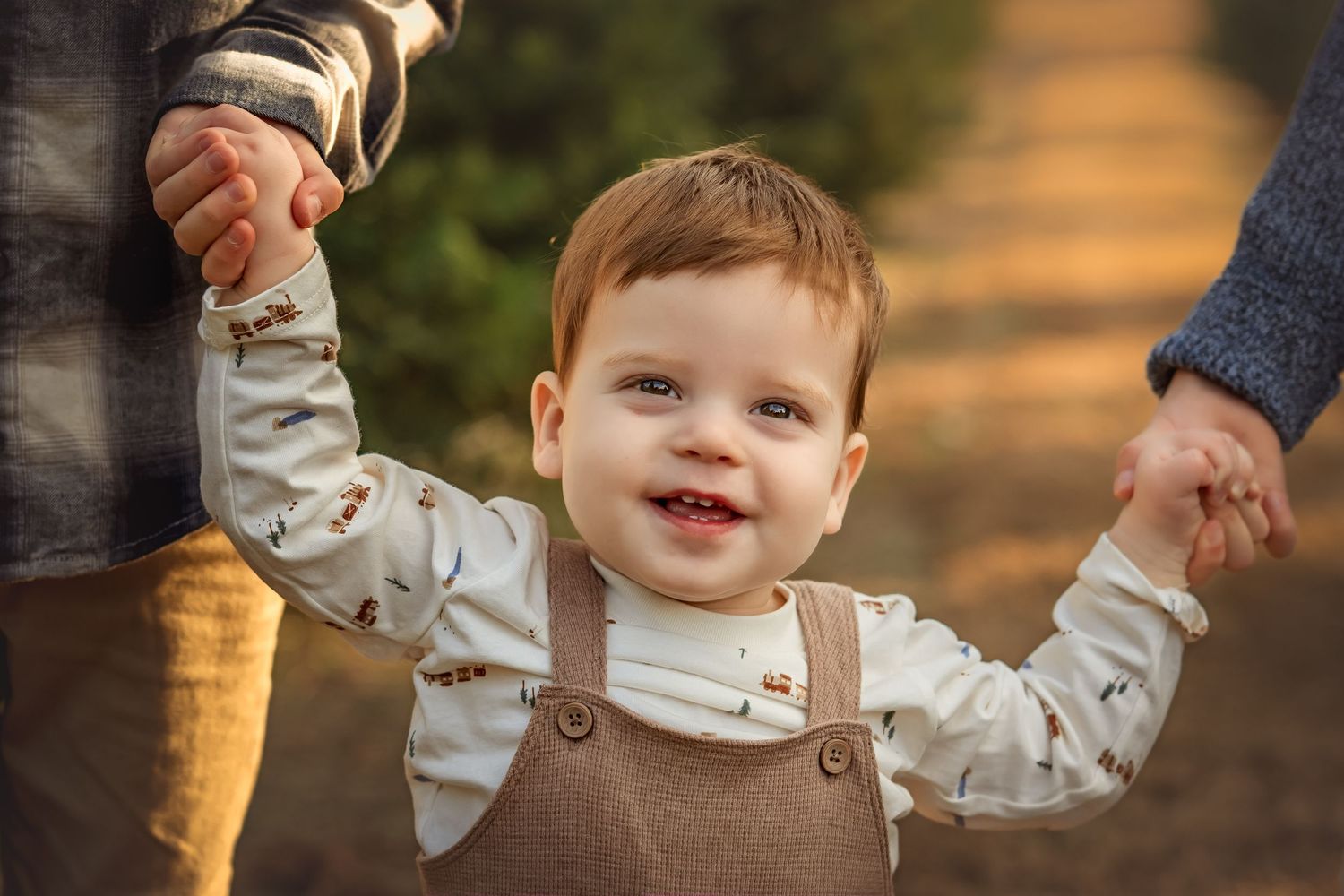 Smiling toddler boy in brown overalls holds hands with two adults while walking outdoors in warm autumn light.