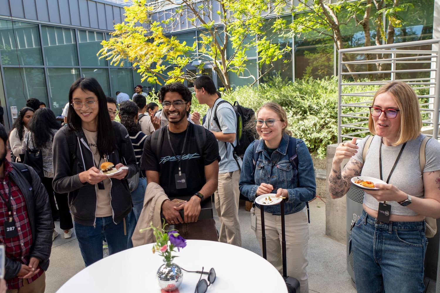 Students smile and socialize outdoors at a campus event, holding plates of food near a white round table.