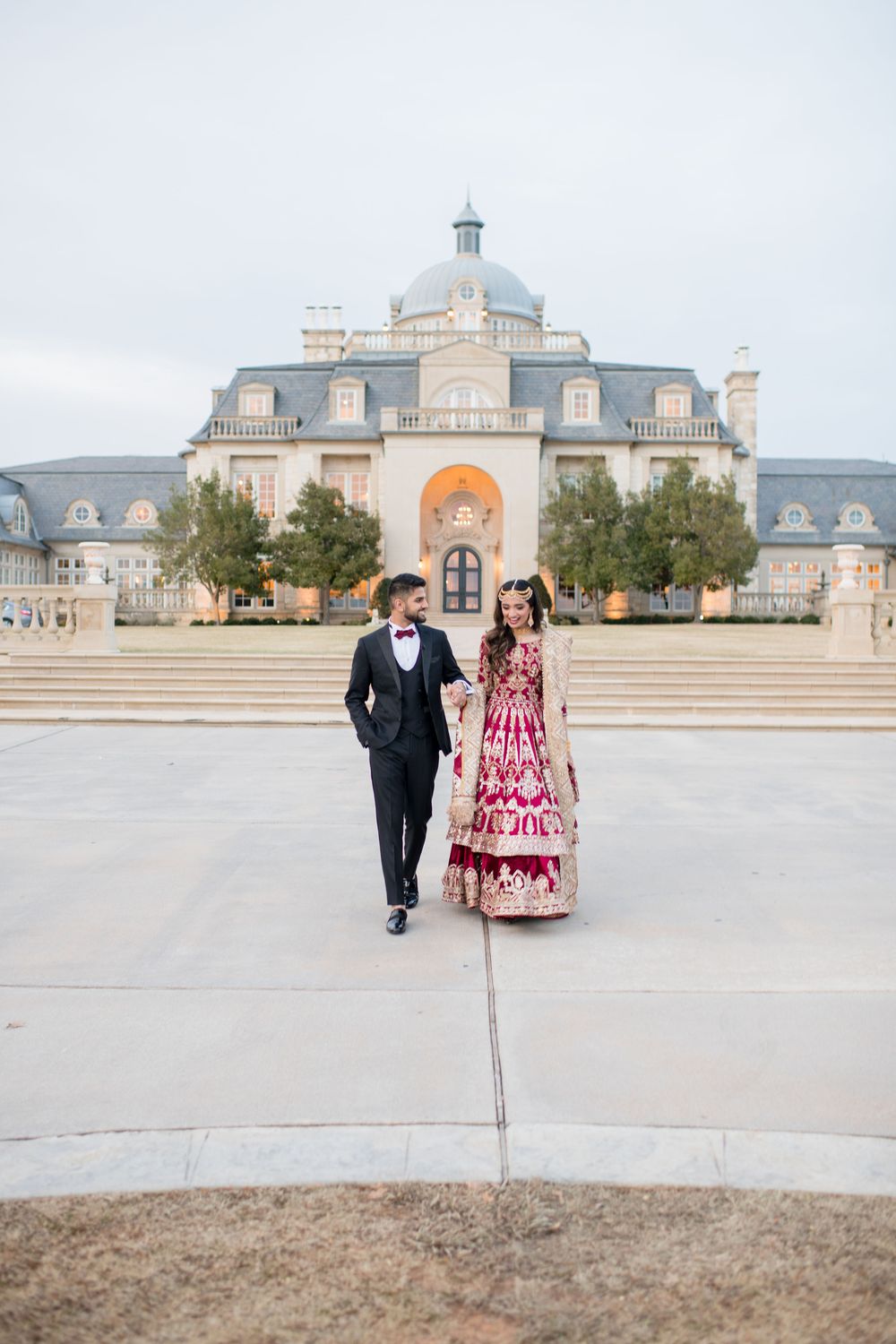 Couple in formal attire walking in front of a grand mansion, bride in red lehenga, groom in black tuxedo.