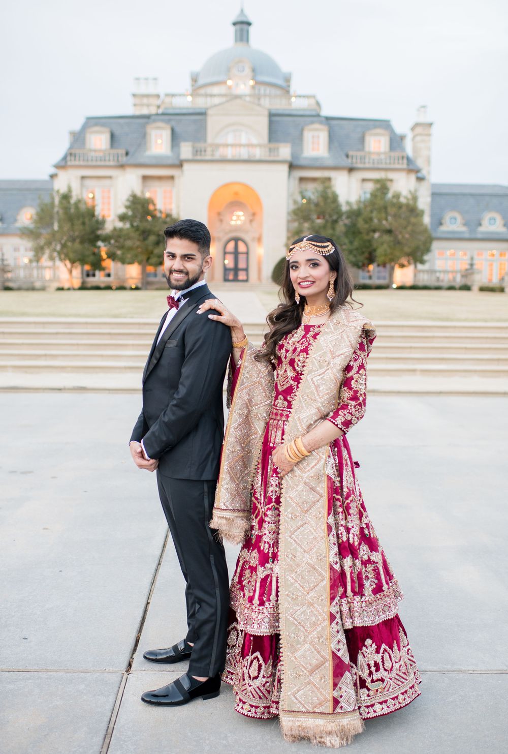 South Asian bride and groom posing in traditional wedding attire outside a grand mansion venue.