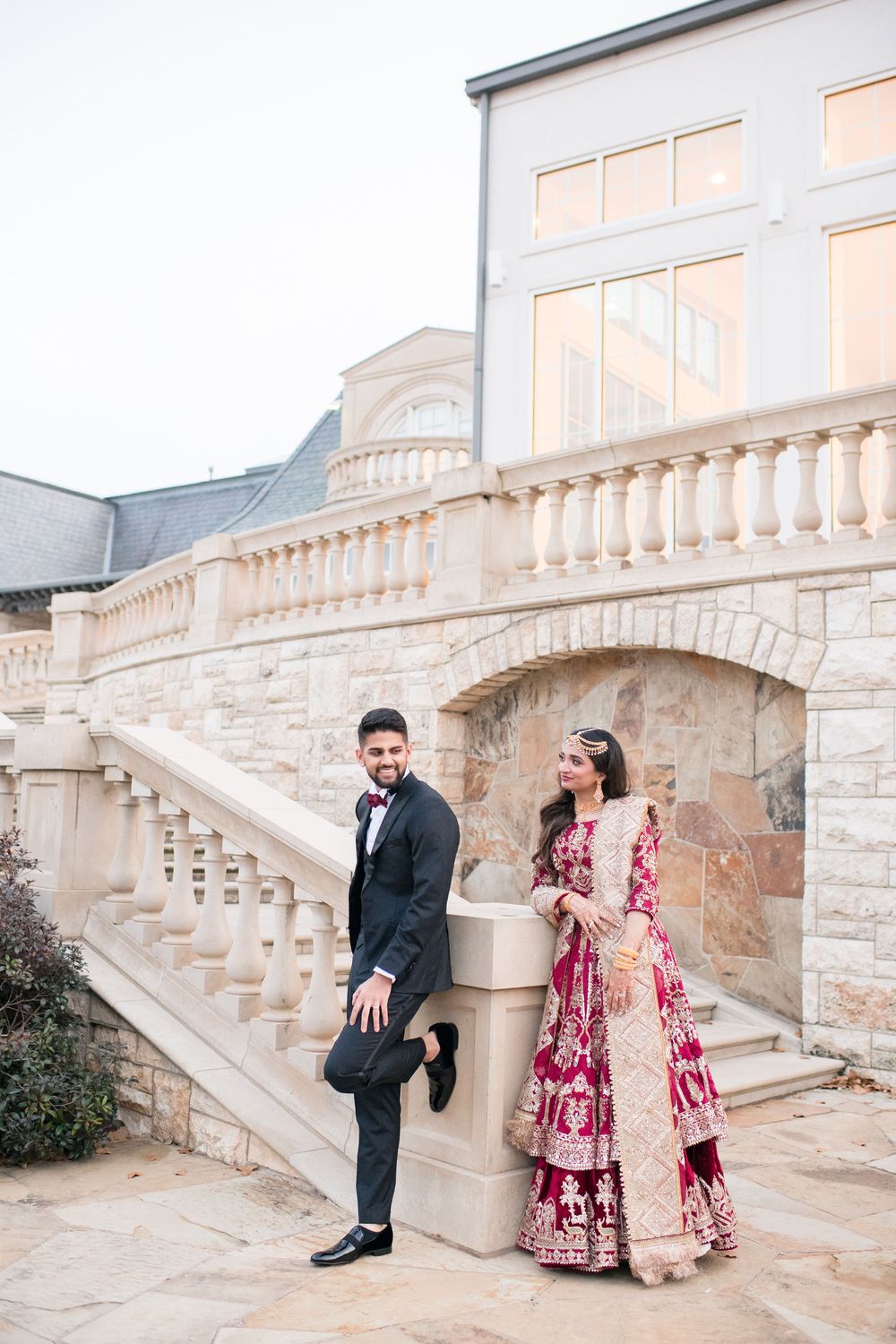 A couple in traditional South Asian wedding attire poses elegantly on stone steps outside a grand venue.