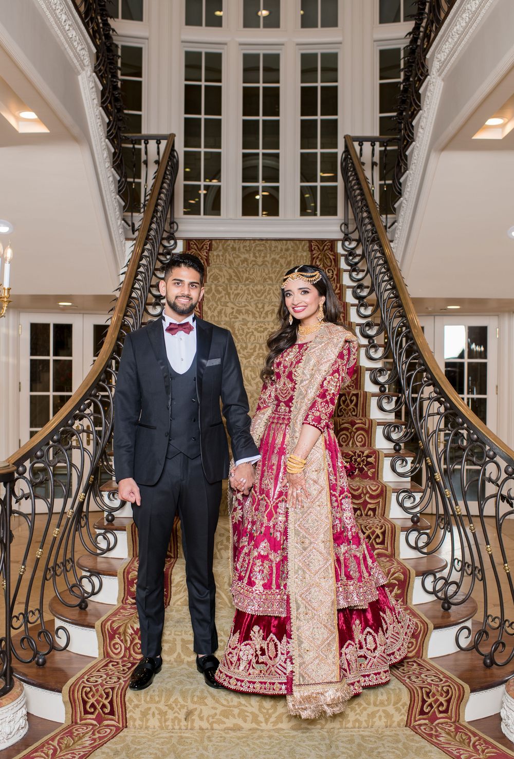 Indian wedding couple in traditional red bridal lehenga and navy suit posing on grand staircase.