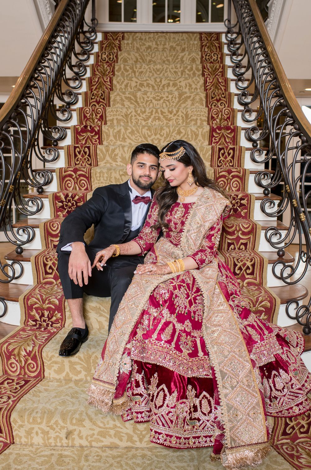 A bride in a red and gold lehenga and groom in a black tuxedo pose on an ornate staircase for their wedding.