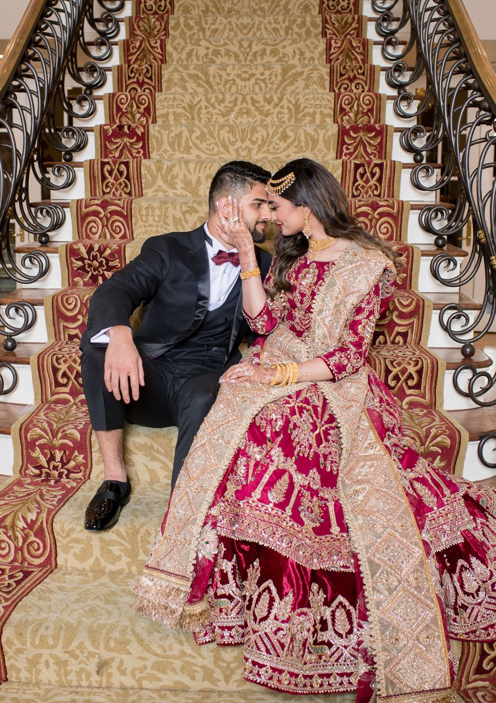 Couple in traditional South Asian wedding attire sit on ornate staircase, bride in red lehenga, groom in black suit.