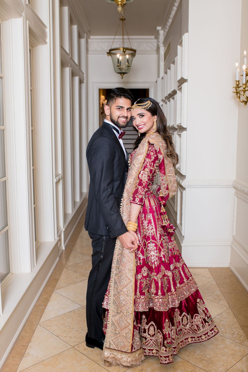 South Asian bride in embroidered red lehenga and groom in black suit pose in elegant white hallway.