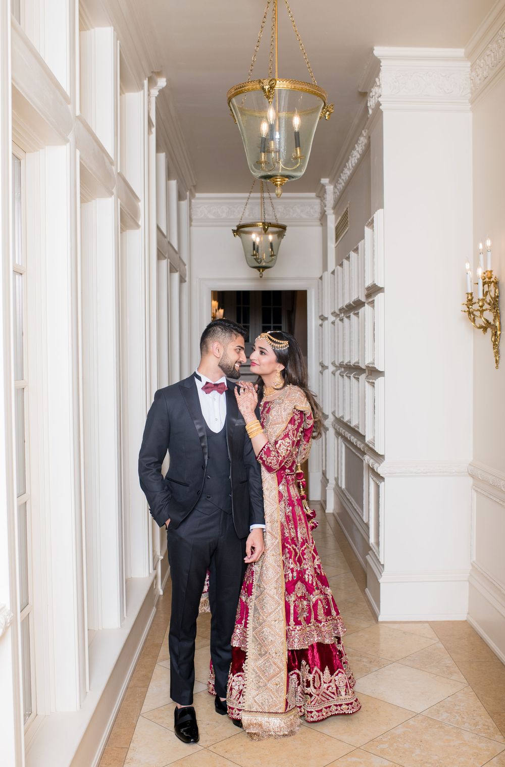 Indian bride and groom share intimate moment in white corridor with gold chandeliers, bride in red lehenga.