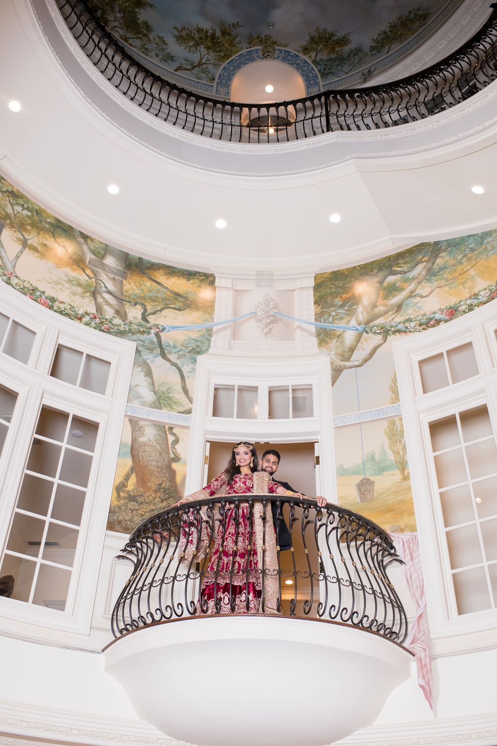 Couple poses on ornate balcony in grand rotunda with scenic murals and elegant dome ceiling.