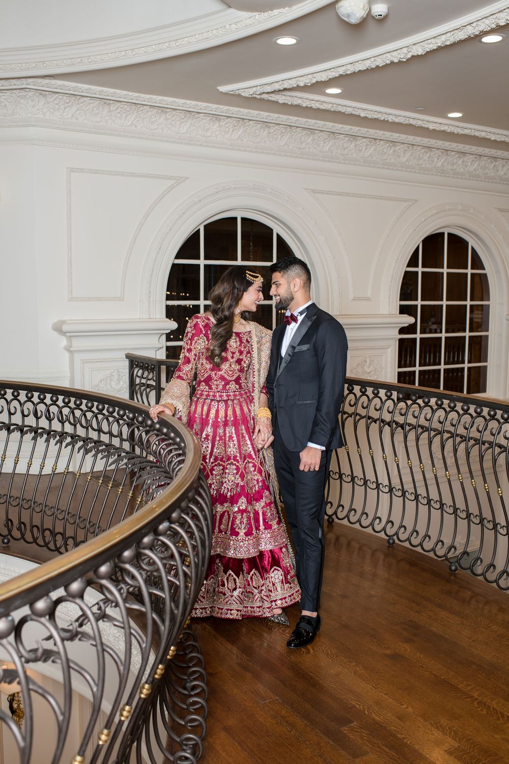 Bride in red lehenga and groom in suit share romantic moment on curved staircase balcony.