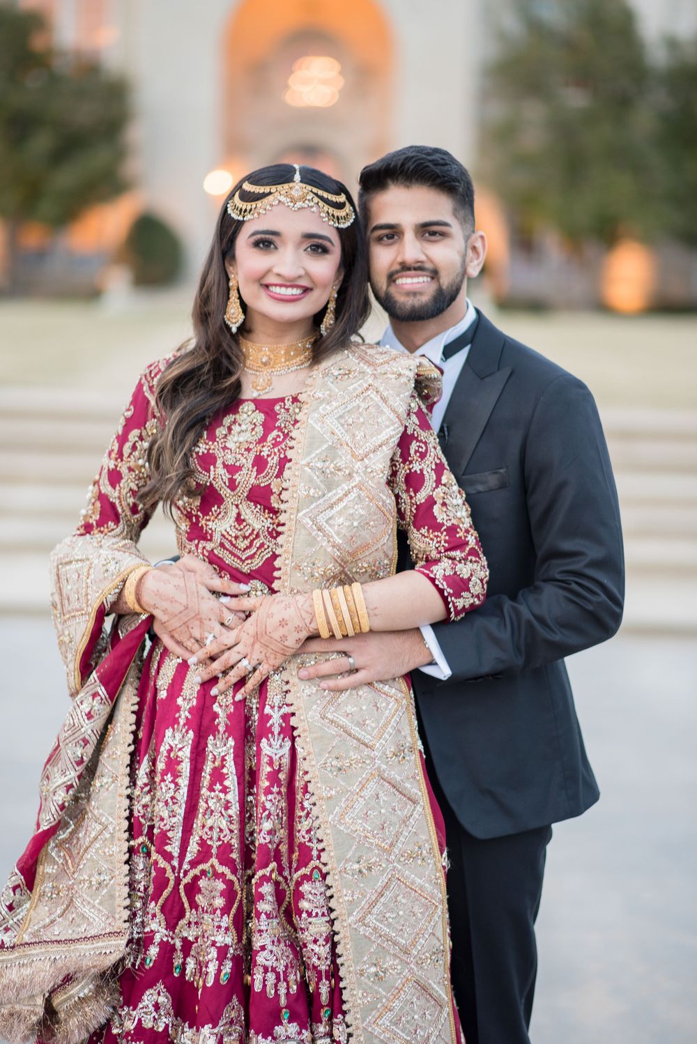 South Asian bride in red and gold lehenga with groom in black tuxedo posing together at wedding.