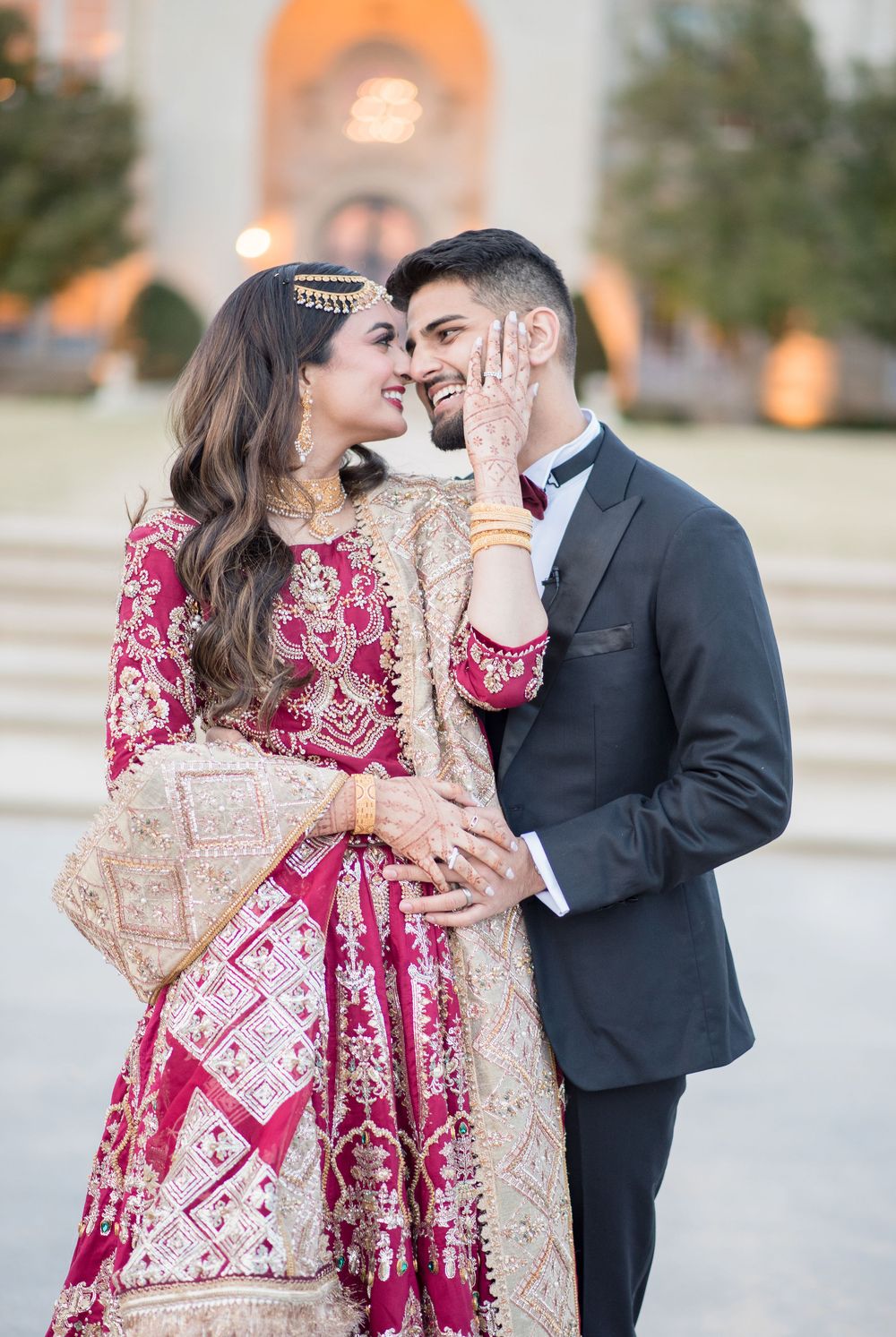 Couple in traditional South Asian wedding attire smiling closely, bride in red lehenga, groom in dark suit.