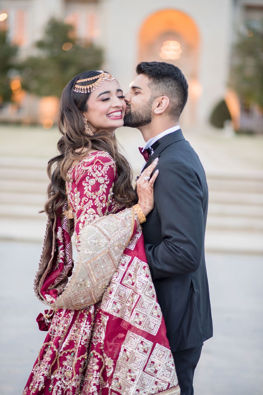 Groom in black tuxedo kissing bride in red gold lehenga on cheek during South Asian wedding celebration.