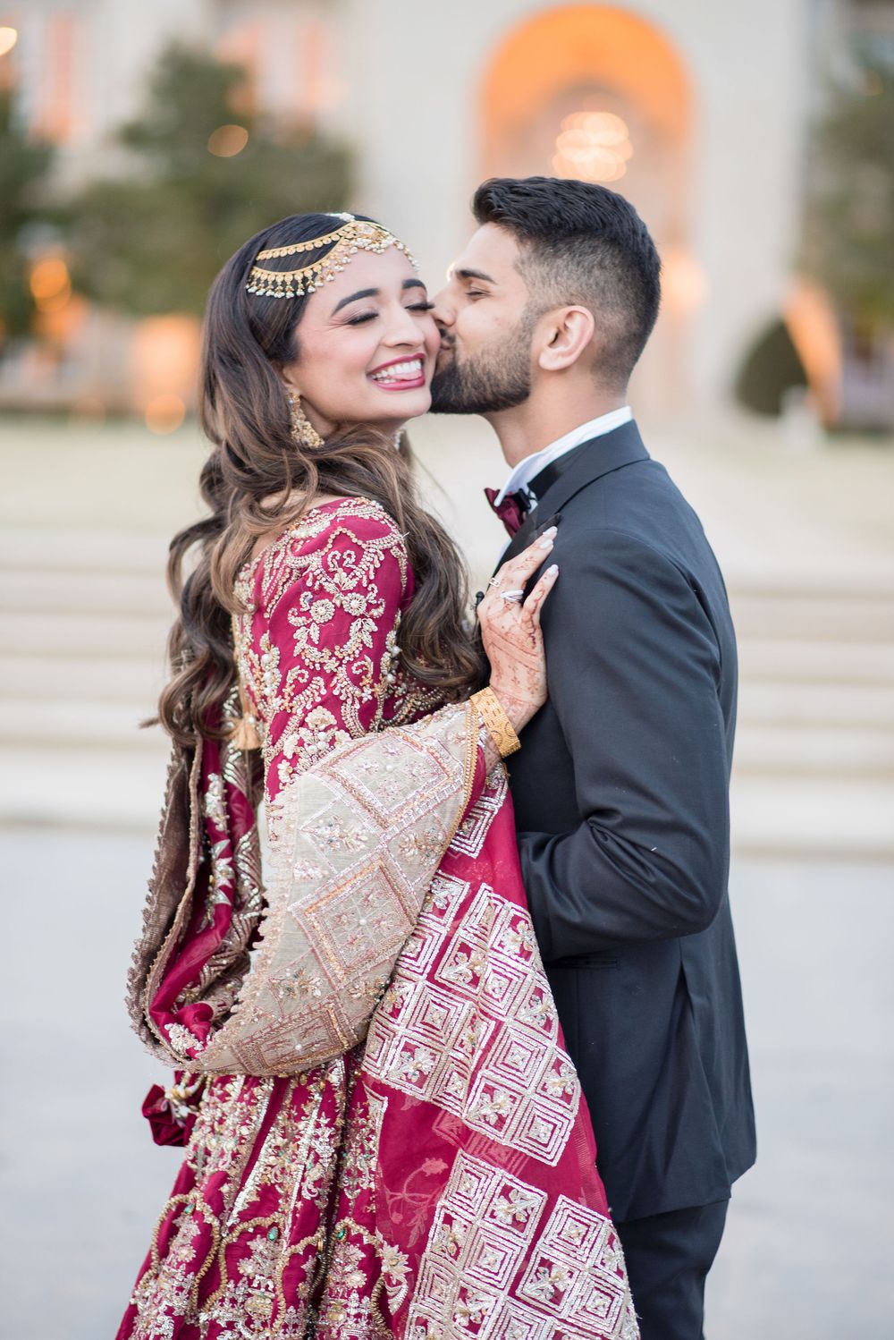 Groom in black suit kisses bride wearing red embroidered lehenga and gold maang tikka at outdoor wedding venue.