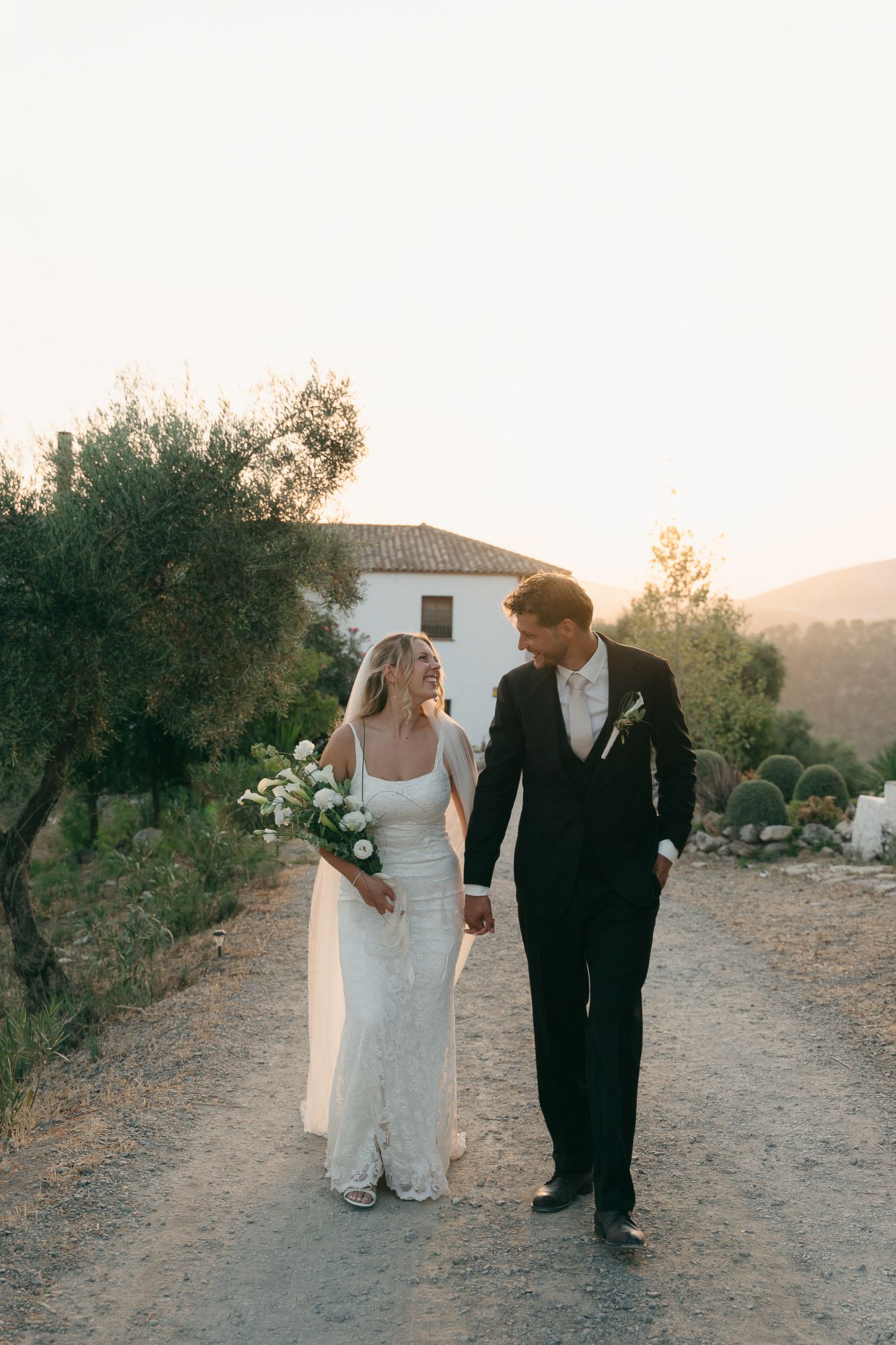 Bride and groom walk hand in hand at golden hour near a white villa surrounded by olive trees.