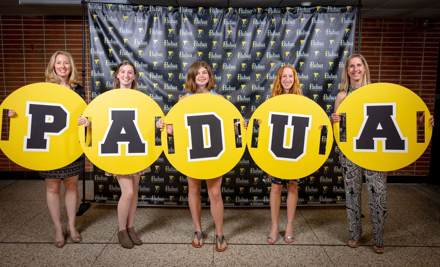 Five women holding large yellow circular letters spelling PADUA pose in front of a branded step-and-repeat backdrop.