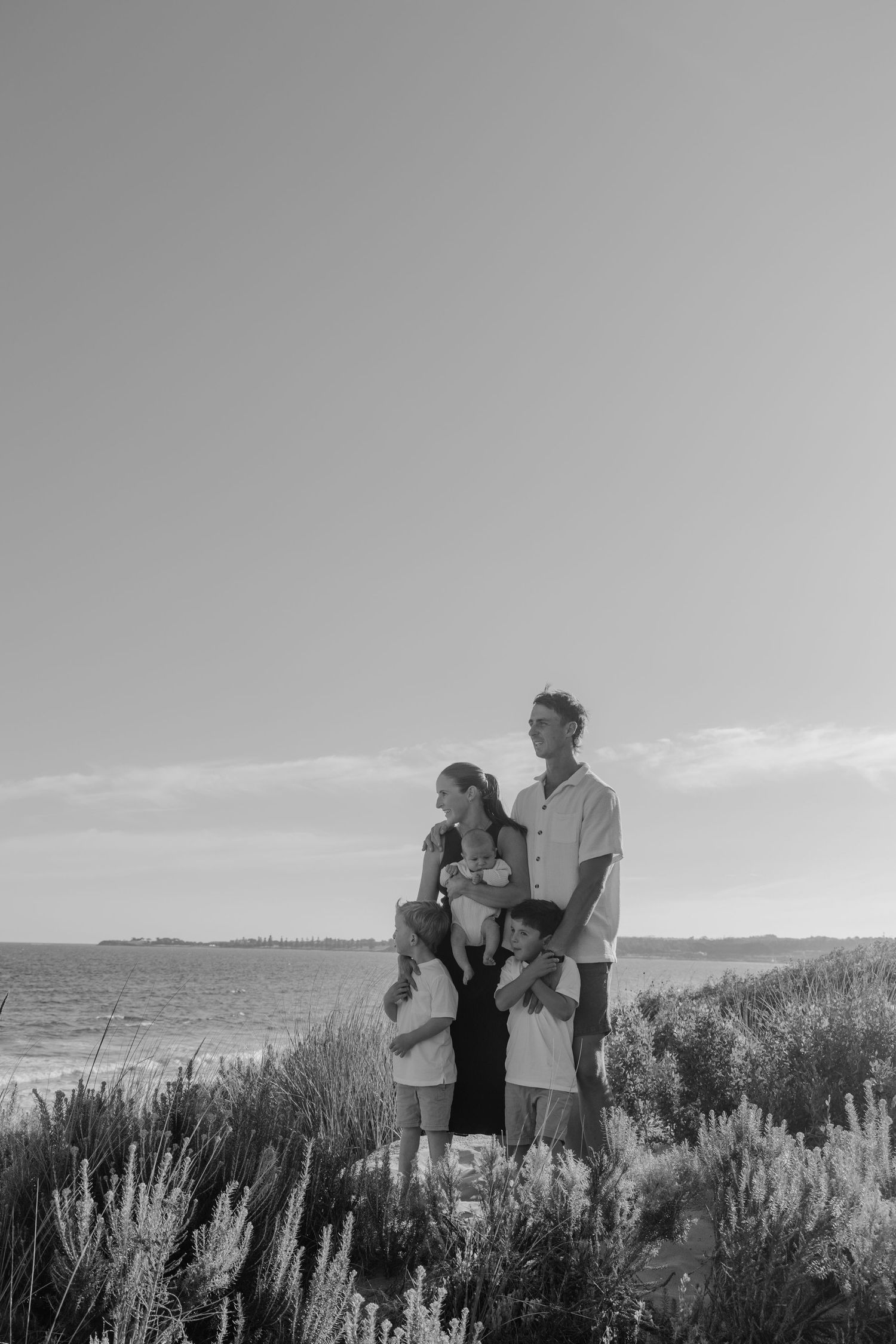 Black and white family portrait on coastal dunes with ocean backdrop, parents holding children among wild shrubs.