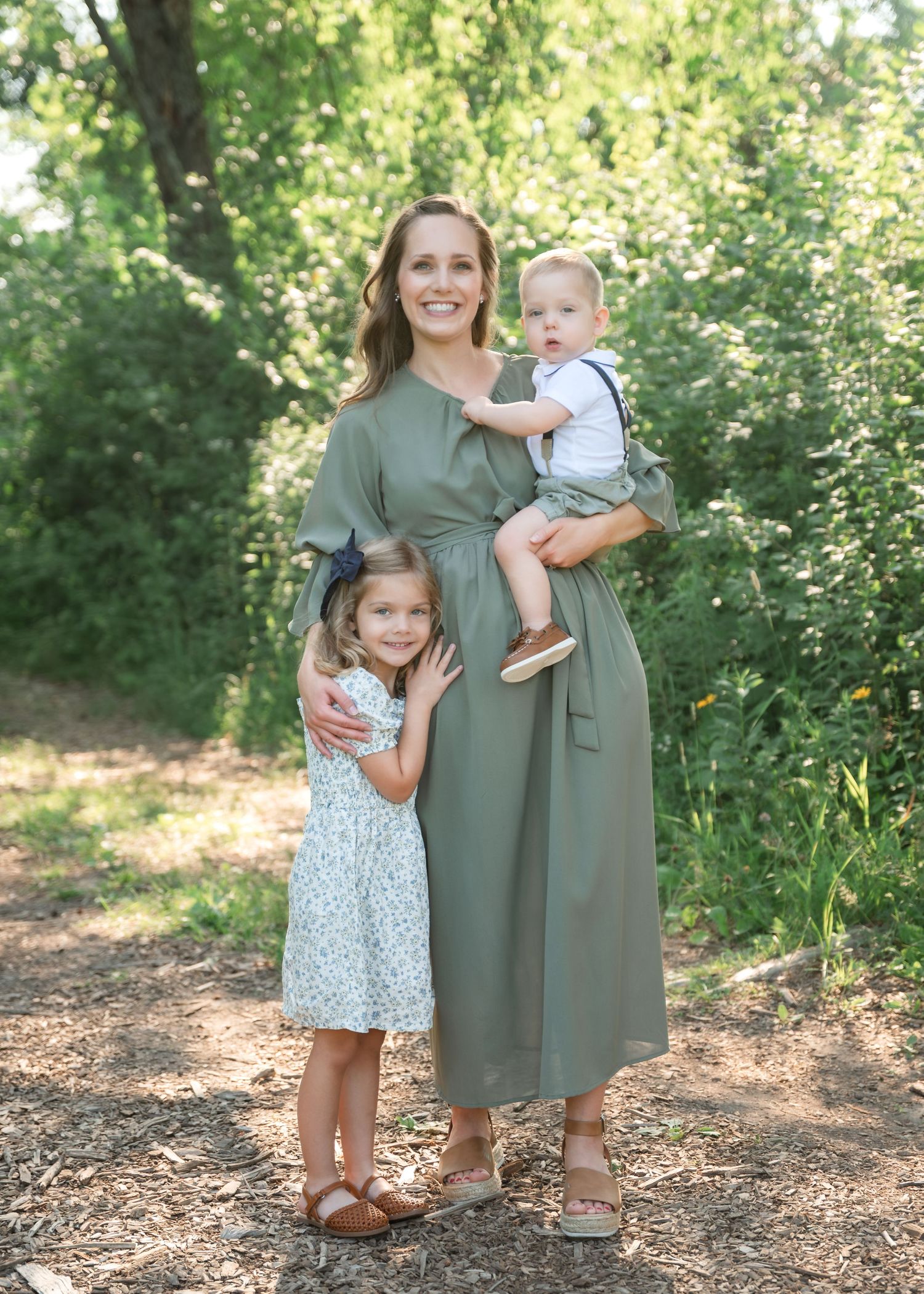 A smiling woman in a green midi dress holds a baby boy while a young girl in a floral dress hugs her side outdoors.