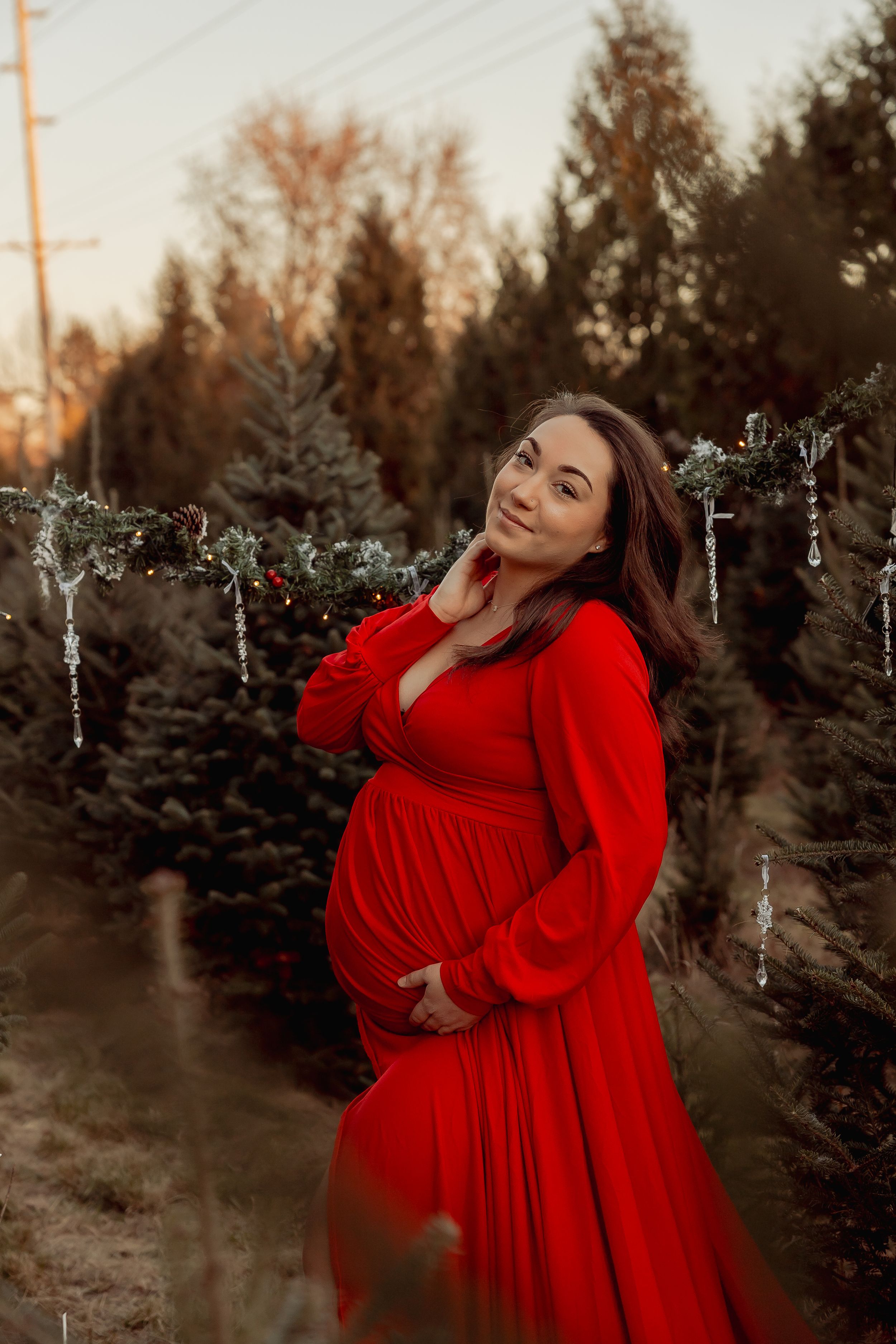 Pregnant woman in red maternity gown poses outdoors among decorated Christmas trees at golden hour.