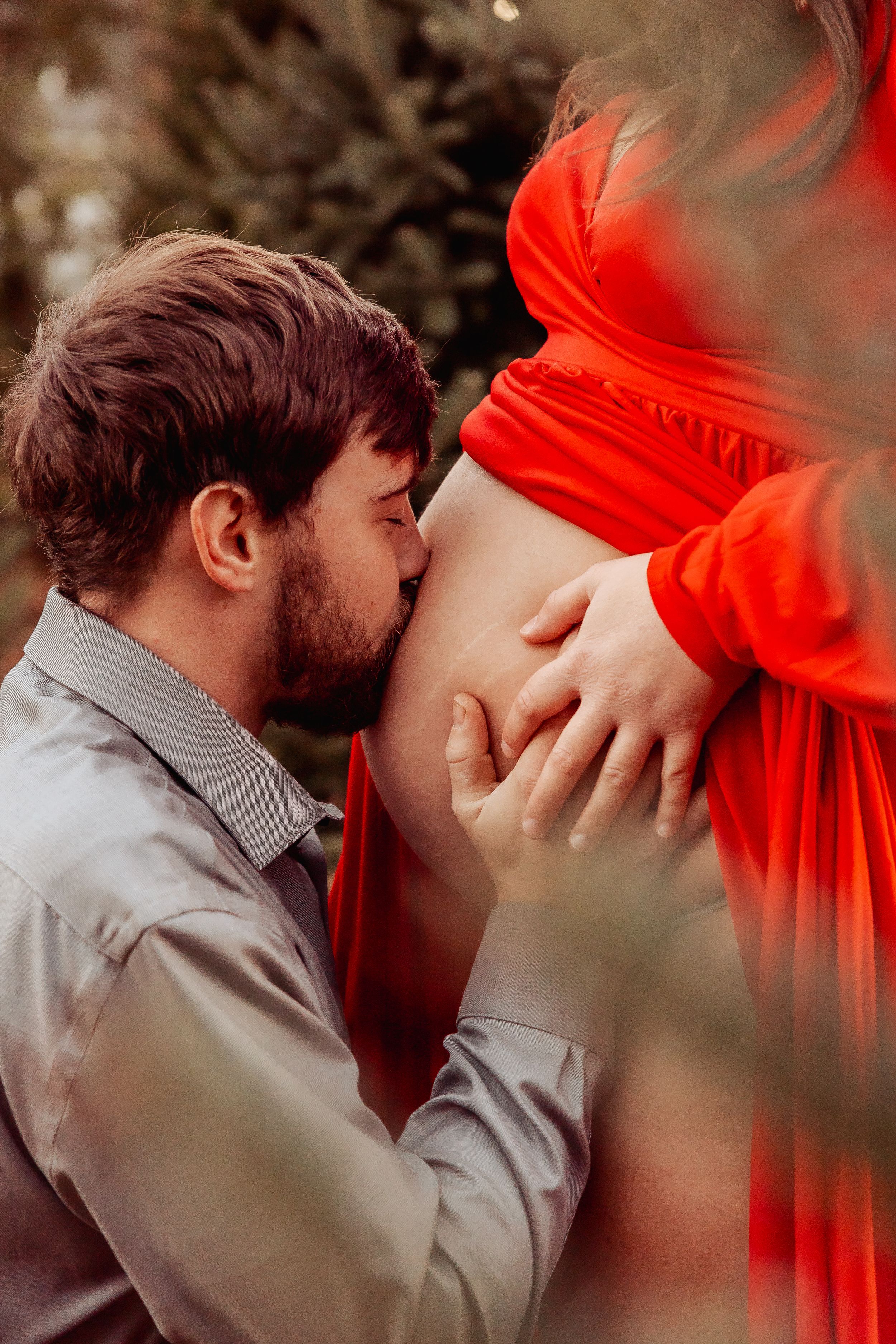 A man tenderly kisses a pregnant woman's bare belly outdoors, as she wears a flowing red dress.