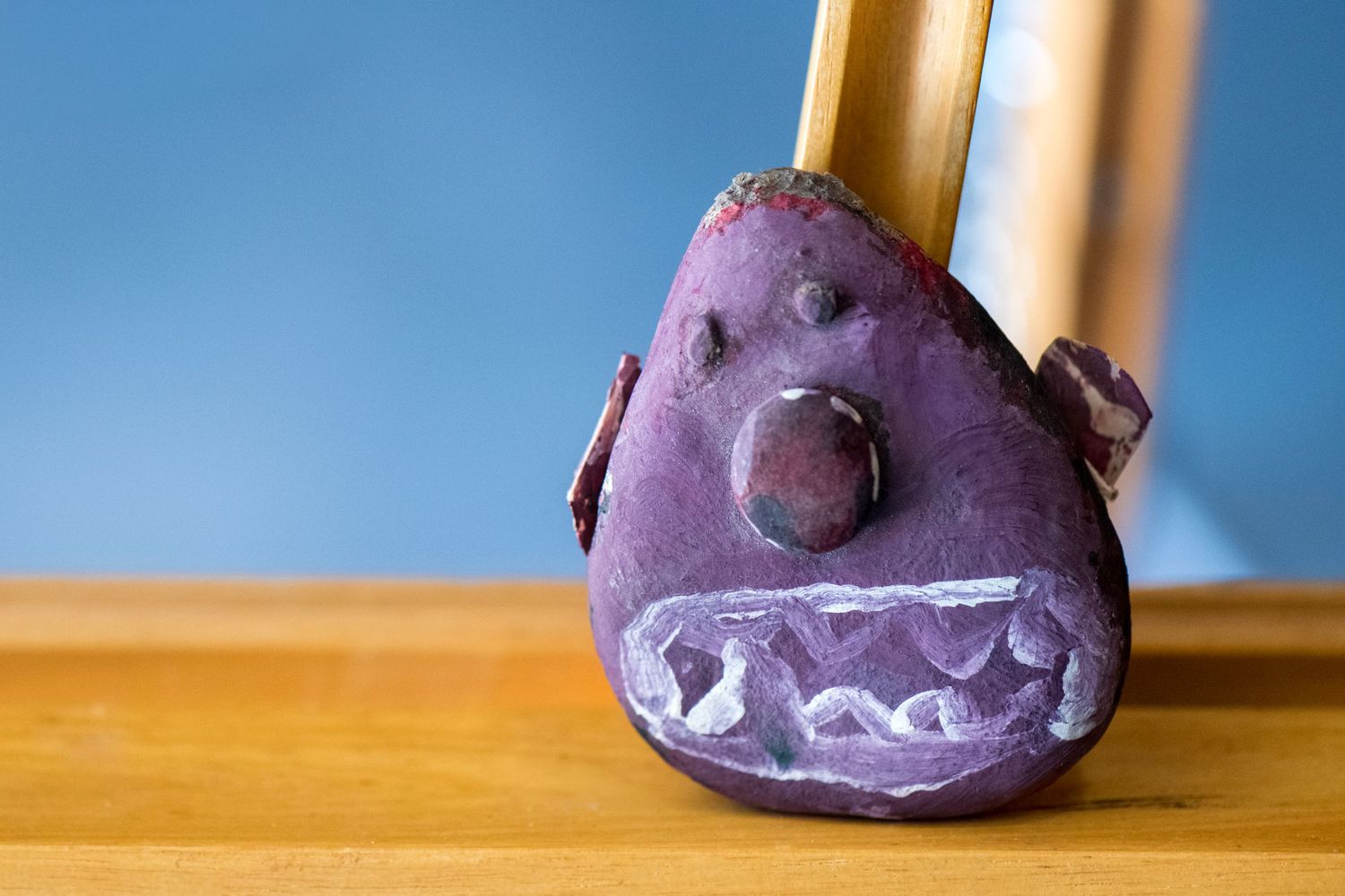 Purple clay monster figurine with white teeth and stick handle displayed on wooden surface against blue background.