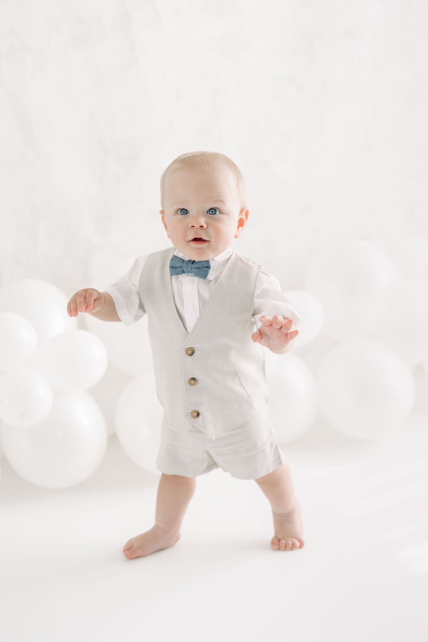 Bébé de 12 mois marchant pieds nus dans un décor lumineux de ballons blancs perlés portant un habit de lin beige de la garde-robe studio