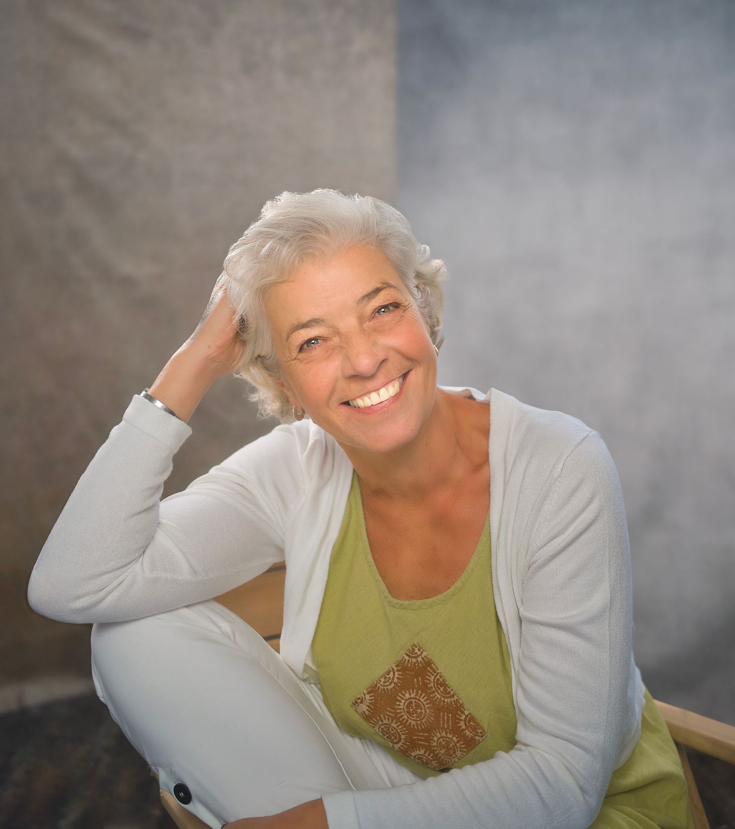 Smiling older woman with short gray hair, wearing a green top and white cardigan, sitting relaxed in a chair.