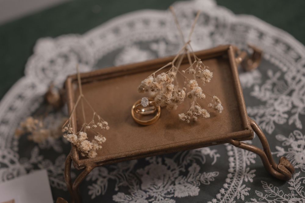 Gold wedding rings on a rustic wooden tray with dried baby's breath flowers, placed on a lace doily.