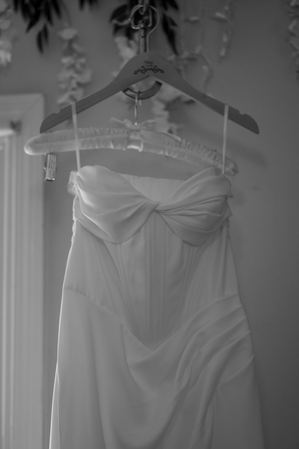 A strapless white wedding dress with a bow detail hangs on a hanger in black and white photo.