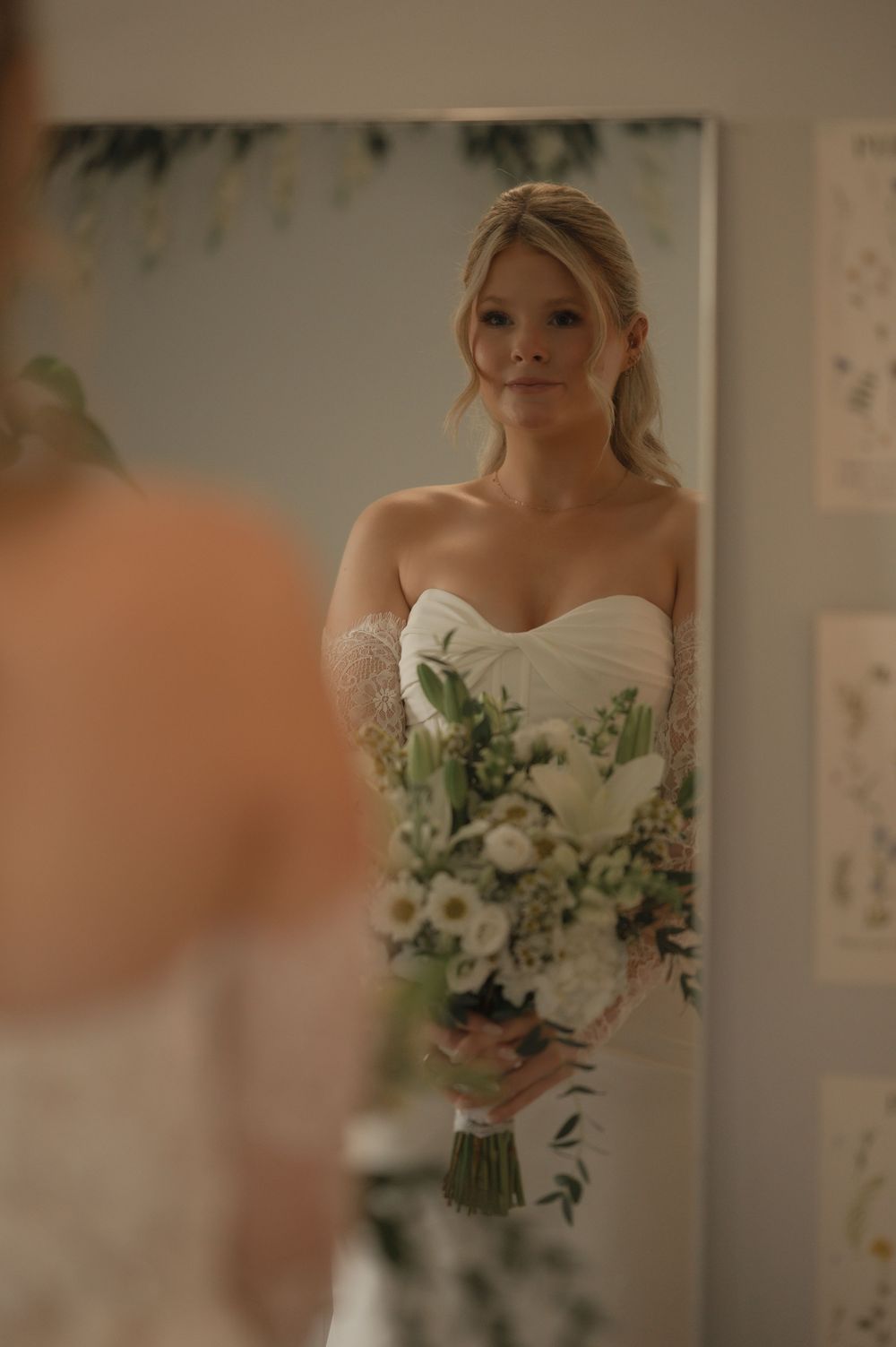 Bride in strapless white gown holds bouquet of white flowers while gazing at her reflection in a mirror.